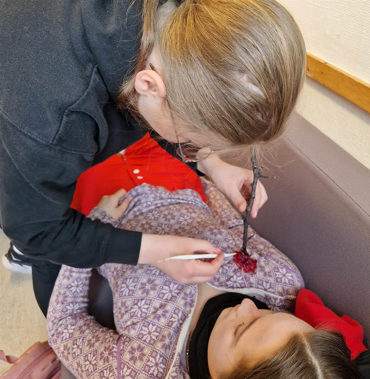 En person legger rød sårsminke rundt en kvist som står ut av brystkassen. til en jenter som ligger.  (Foto) - Klikk for stort bilde